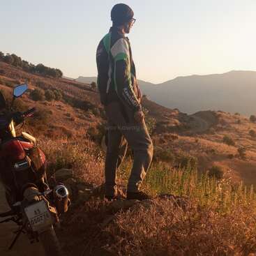 Un homme se tient sur une colline, contemplant les montagnes, vêtu d'un blouson et d'un bonnet Kawasaki, avec une moto garée à proximité au premier plan.