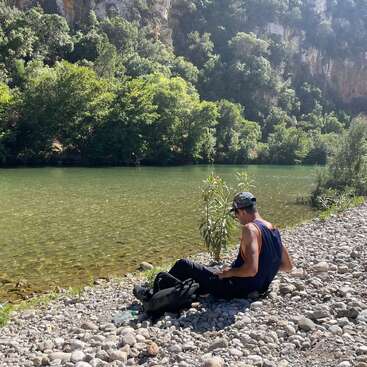 Ein Mann sitzt an einem felsigen Flussufer, umgeben von Grün und Bäumen, und genießt das Sonnenlicht. Im Hintergrund erheben sich Berge, die eine friedliche, natürliche Szene schaffen.