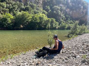 A man sits on a rocky riverbank, surrounded by greenery and trees, enjoying the sunlight. Mountains rise in the background, creating a peaceful, natural scene.