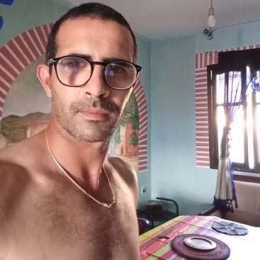 Un homme aux cheveux courts et portant des lunettes, torse nu, se tient debout dans une pièce colorée avec des arches peintes, une nappe rayée et la lumière du soleil qui pénètre par la fenêtre.