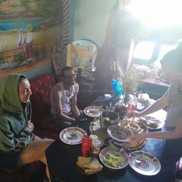 A imagem mostra uma mulher servindo comida para duas pessoas sentadas em uma mesa, com um mural na parede e uma janela ao fundo, evocando um ambiente aconchegante.