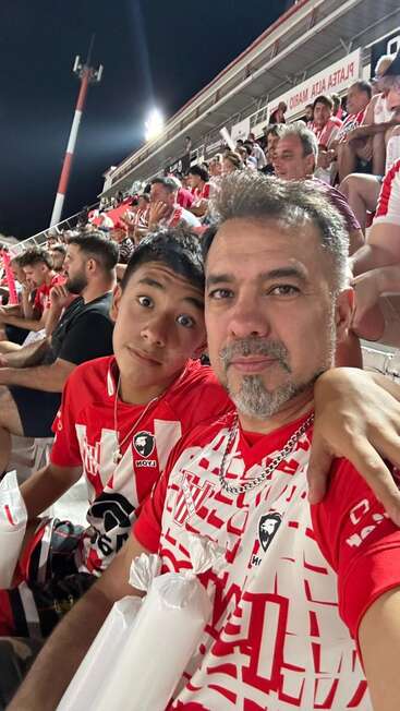 A father and son take a selfie at a crowded soccer stadium. Both wear red and white team jerseys. The stands are filled with cheering fans.