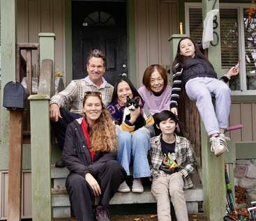 Une famille multigénérationnelle est assise sous le porche, souriante. Il y a des adultes, des enfants, une femme âgée et un chat, qui profitent d'un moment agréable.