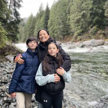 A woman and two children, all wearing jackets, stand on a rocky riverside in a forest, smiling together. Tall green trees line the background.