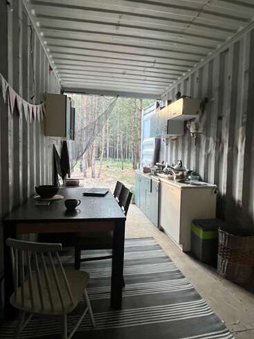 Dentro de un contenedor de transporte reutilizado, una acogedora cocina y un comedor con decoración rústica, muebles sencillos y vistas al bosque a través de una puerta abierta, invitan a la naturaleza a entrar.