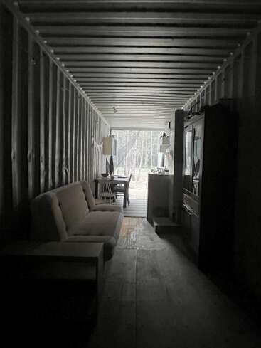 Esta imagen muestra un contenedor de transporte poco iluminado convertido en un espacio habitable, con un sofá, una mesa, una silla, un armario y una ventana que deja ver árboles en el exterior.