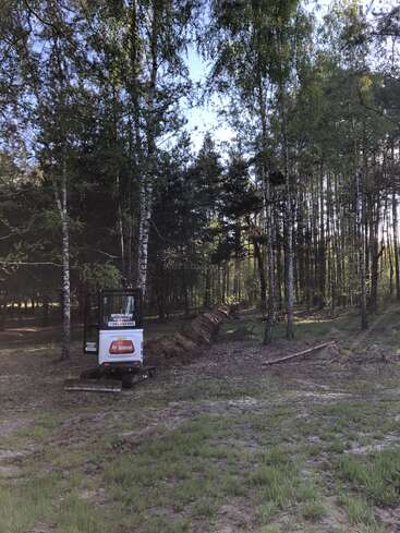A small Bobcat excavator sits at the edge of a forest, next to a freshly dug trench extending through the trees, under clear, sunny skies.