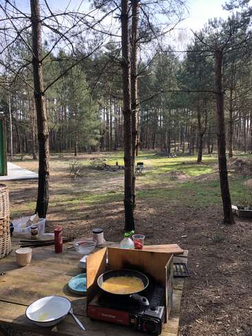 A rustic outdoor kitchen setup in a forest with a frying pan on a portable stove, surrounded by cooking supplies on a wooden picnic table under tall pine trees.