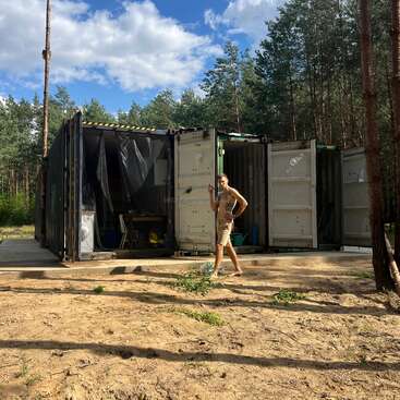 A person stands outside converted shipping containers in a forest clearing. The containers’ doors are open, revealing a workspace inside. Sunlight filters through tall trees nearby.