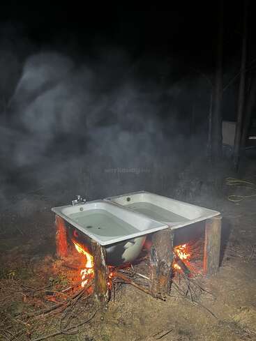 Two bathtubs are outdoors at night, each filled with water, elevated on wooden logs, and heated by a fire burning beneath them. Smoke surrounds everything.
