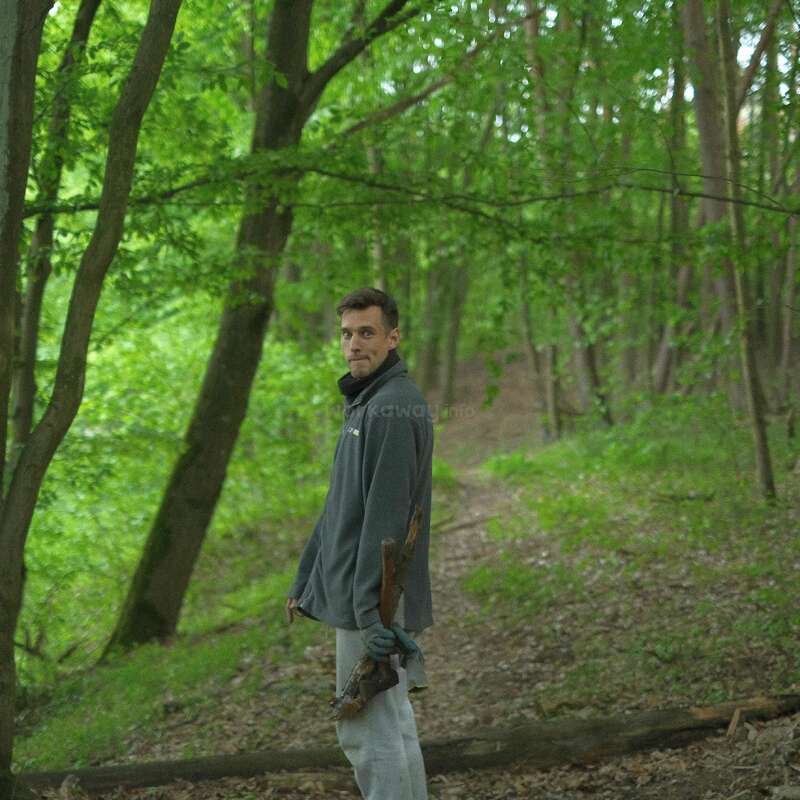 Un hombre está de pie en un frondoso bosque verde, sujetando una rama, vestido con ropa informal y mirando hacia atrás por encima del hombro en un sombreado camino forestal.