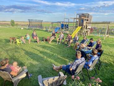 Un groupe de personnes est assis en cercle sur des chaises de jardin dans un jardin, avec des enfants, des chiens et une aire de jeux en arrière-plan, profitant du beau temps.