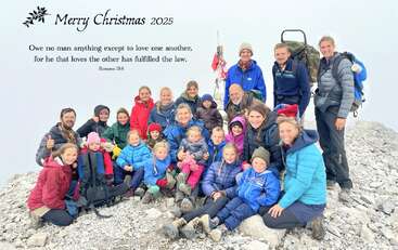 Une grande famille, vêtue de tenues d'hiver, pose au sommet d'une montagne rocheuse. L'image comporte un message de vœux de Noël et un verset biblique sur l'amour.