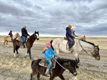 Sous un ciel nuageux, un groupe d'enfants et d'adultes chevauchent des chevaux et un poney sur un chemin de terre à la campagne, au milieu de champs à perte de vue et d'herbe sèche.