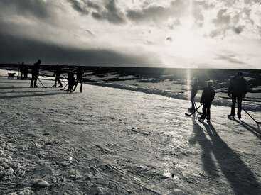 L'image représente un groupe de personnes jouant au hockey sur un lac gelé, le soleil traversant les nuages et jetant une lueur chaleureuse sur la scène.