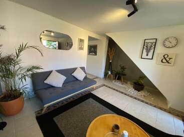 Este salón cuenta con un moderno sofá gris con cojines blancos, arte en la pared, un espejo redondo, plantas, un reloj y una decoración acogedora y minimalista.