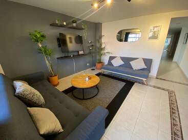 Un moderno salón cuenta con dos sofás, una mesa de centro redonda, televisión, plantas, colores neutros, estanterías de pared, un gran espejo y cojines decorativos que aumentan la calidez.