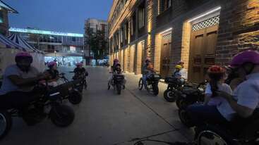 Un groupe de personnes portant des casques roulent sur des tricycles électriques au crépuscule près d'un bâtiment en briques bien éclairé. La zone environnante semble animée et urbaine.