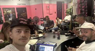 Un groupe de personnes est assis autour d'une table dans une pièce moderne au thème rose, en train de discuter. Des ordinateurs portables, des tasses de café et des blocs-notes sont visibles sur la table.