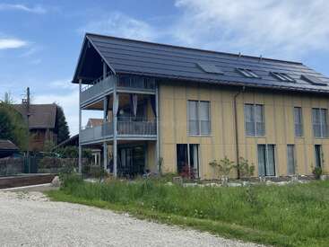 Uma casa moderna de madeira com grandes painéis solares no telhado, duas varandas e muitas janelas, cercada por grama verde sob um céu azul.