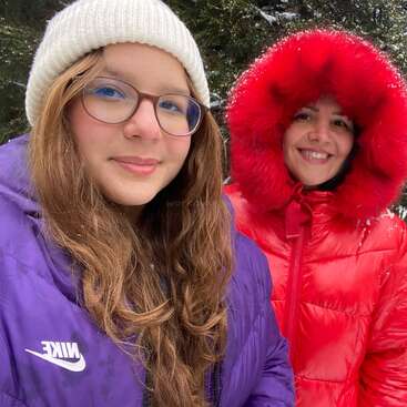 Zwei Menschen in Winterkleidung, die lächelnd im Freien stehen. Einer trägt eine lila Nike-Jacke und eine Brille, der andere einen roten, pelzgefütterten Parka. Schnee und Bäume sind zu sehen.