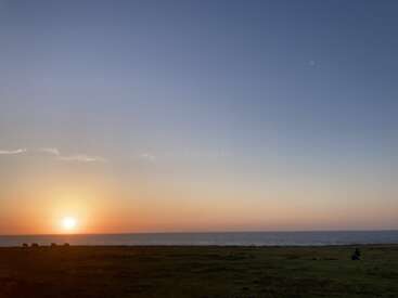 Una serena puesta de sol sobre el océano, con el sol cerca del horizonte, proyectando cálidos colores sobre el cielo. Mar en calma, primer plano cubierto de hierba y una figura lejana.
