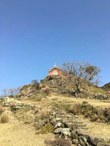 Una pequeña iglesia con cruces se alza sobre una colina rocosa, rodeada de árboles desnudos, escalones de piedra, hierba seca y un cielo azul despejado.