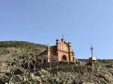 Una iglesia de ladrillo rojo se alza sobre una colina rocosa, rodeada de árboles desnudos y muros de piedra, bajo un cielo azul despejado y un fondo de montañas verdes y boscosas. Ambiente apacible.