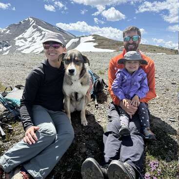 Una familia de tres miembros y su perro están sentados al aire libre sobre un terreno rocoso, con montañas nevadas y un cielo azul al fondo; todos sonríen y disfrutan del sol.