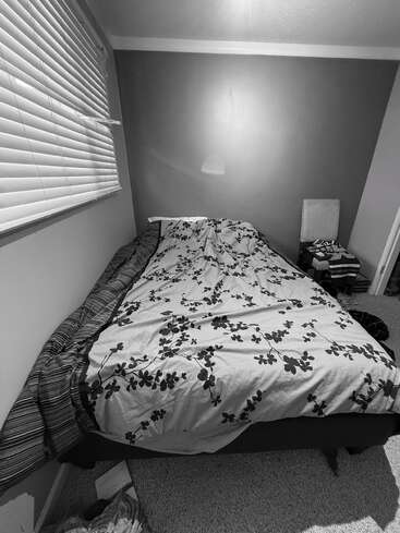 Esta fotografía en blanco y negro muestra una cama bien hecha con una manta de flores, junto a una ventana con persianas, y una silla con ropa encima.