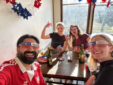 Quatre amis portant des lunettes de soleil aux couleurs du drapeau américain jouent aux cartes autour d'une table, tout en dégustant des boissons et des amuse-bouches, et font la fête ensemble dans une pièce baignée de soleil et décorée de manière festive.