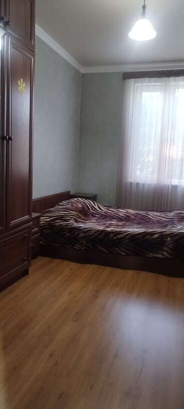 Esta imagem mostra um quarto arrumado com piso de madeira, um guarda-roupa grande, uma cama com um cobertor estampado, paredes claras e uma janela com cortinas transparentes.