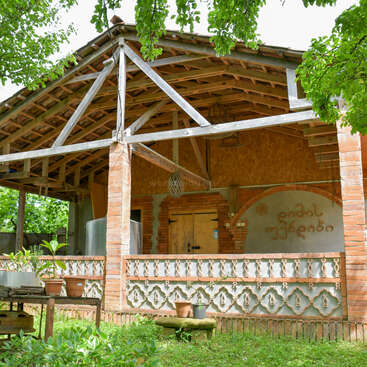 Uma casa rústica de tijolos com uma varanda aberta, cercada por árvores frondosas. Vasos de plantas decoram a varanda e a mesa, criando um ambiente aconchegante e natural.