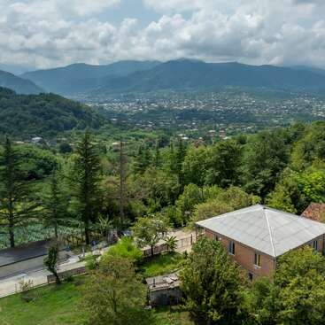 Uma paisagem pitoresca com uma casa cercada por árvores verdejantes, com vista para um vale repleto de edifícios. No fundo, avistam-se montanhas e um céu nublado.
