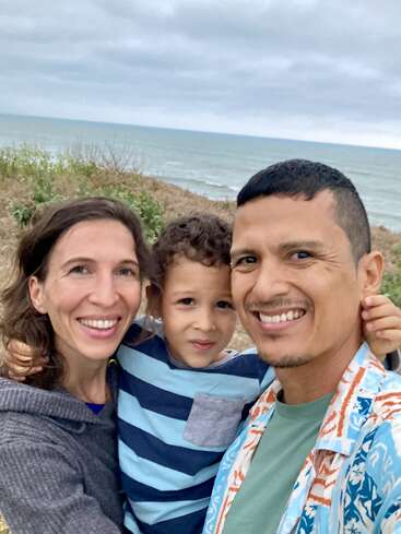 Une famille heureuse de trois personnes pose ensemble pour un selfie au bord de la mer. Le ciel est nuageux et les vagues de l'océan sont visibles en arrière-plan.
