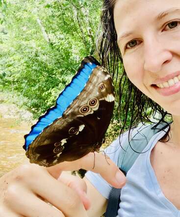 Une femme souriante aux cheveux mouillés tient un grand papillon bleu et brun sur son doigt, entourée d'arbres verdoyants dans un cadre extérieur lumineux et naturel.