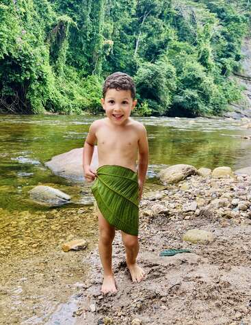 Un jeune garçon souriant se tient au bord d'une rivière et porte une grande feuille verte en guise de jupe. Une forêt luxuriante entoure l'eau, créant une scène ludique et naturelle.