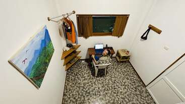 Dans une petite pièce bien rangée, un homme est assis à un bureau et utilise un ordinateur portable. Des vêtements sont accrochés au mur ; un tableau et une fenêtre sont visibles.