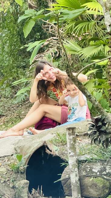 Une femme et un jeune enfant sont assis sur un rocher dans une forêt verdoyante. Ils sourient et font des gestes ludiques de la main, profitant du temps qu'ils passent ensemble en plein air.
