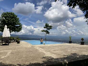 Un homme se tient dans une piscine à débordement sereine surplombant un vaste paysage sous un ciel bleu vif avec des nuages cotonneux, entouré de plantes en pot et d'arbres d'ombrage.