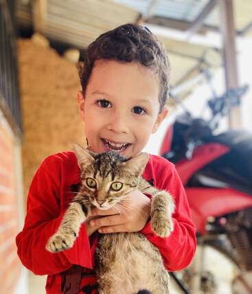 Un jeune garçon souriant, vêtu d'une chemise rouge, tient joyeusement un chat tabby. Ils sont en plein air, près d'une moto, dans une ambiance chaleureuse et joyeuse.