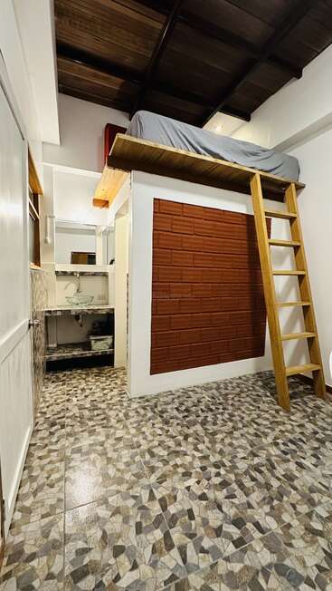 This image shows a small room with a loft bed accessed by a wooden ladder, mosaic tile flooring, a brick accent wall, and a compact bathroom area.