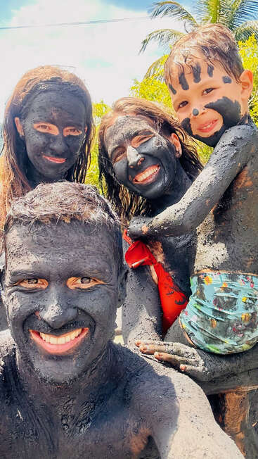 Une famille souriante de quatre personnes est couverte de boue noire, s'amusant en plein air sous un ciel ensoleillé et des palmiers, créant ainsi des souvenirs de vacances joyeux et désordonnés.