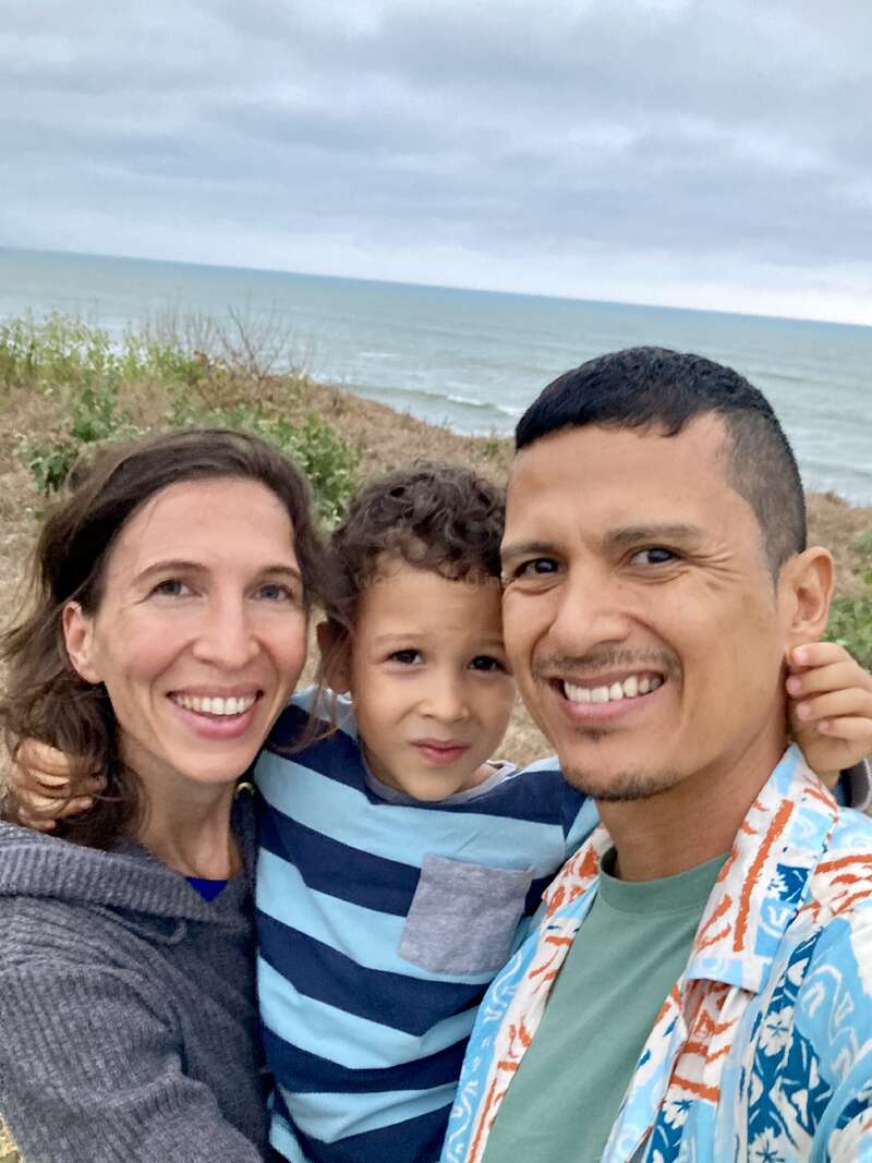 Une famille heureuse de trois personnes pose ensemble pour un selfie au bord de la mer. Le ciel est nuageux et les vagues de l'océan sont visibles en arrière-plan.