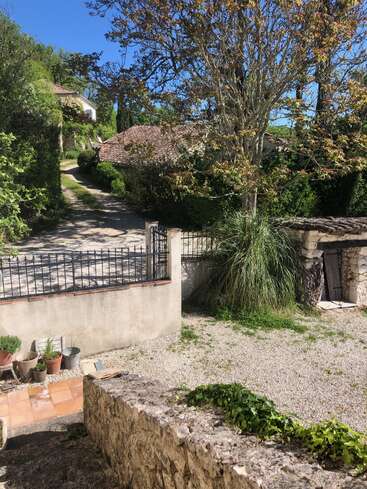 A sunlit courtyard with gravel ground, stone steps, potted plants, and a tree. A path leads past garden walls to rustic tiled-roof houses surrounded by greenery.