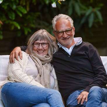 Um casal de idosos sorridentes senta-se bem juntinho em um banco ao ar livre, ambos usando óculos e roupas casuais, parecendo felizes e relaxados, cercados por folhagem verde.