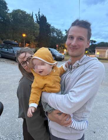 A smiling man holds a happy baby in a yellow hoodie, standing beside a woman with glasses. They are outdoors in a parking lot at dusk.