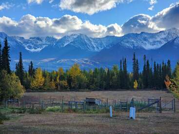 Snow-capped mountains rise majestically behind a dense forest of evergreen and autumn-colored trees, with a fenced grassy field in the foreground under a cloudy sky.