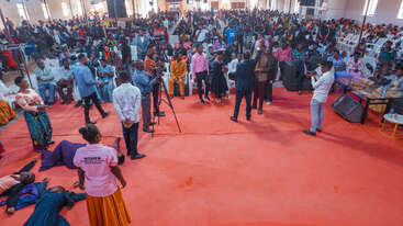 Una gran congregación se reúne en el interior de una iglesia. Varias personas están en el escenario, algunas parecen presentar o filmar. Dos personas están tumbadas en el suelo, ayudadas por un ujier.