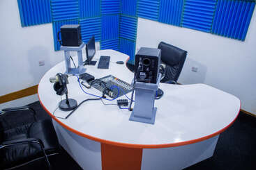 Un moderno estudio de radio o podcast con micrófonos, auriculares, altavoces, mesa de mezclas, ordenador, sillas negras y paneles de espuma acústica azul en las paredes. Un entorno limpio y profesional.